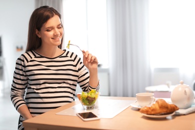 Young pregnant woman eating vegetable salad at table in kitchen