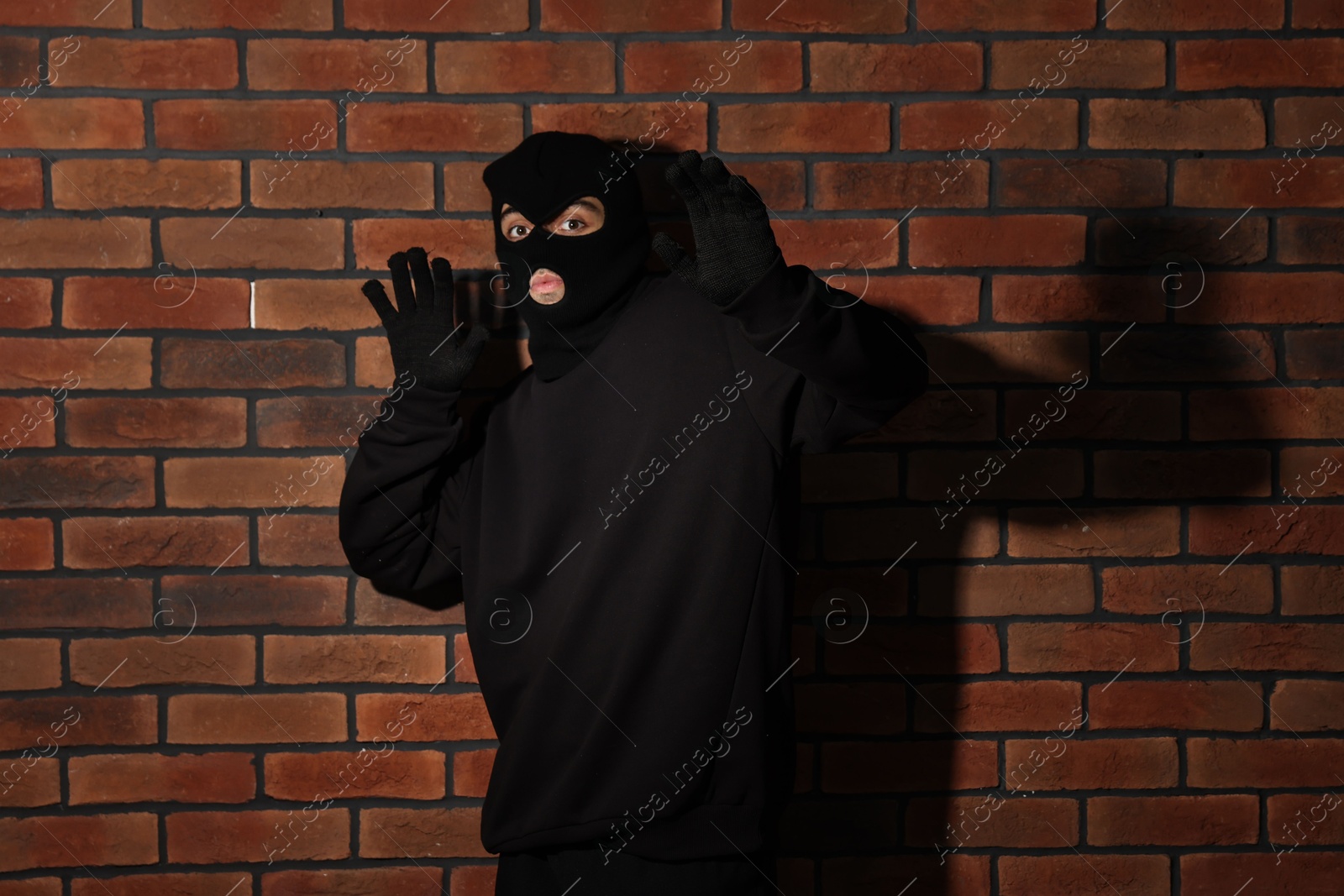 Photo of Thief in balaclava raising hands against red brick wall