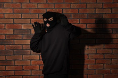 Thief in balaclava raising hands against red brick wall