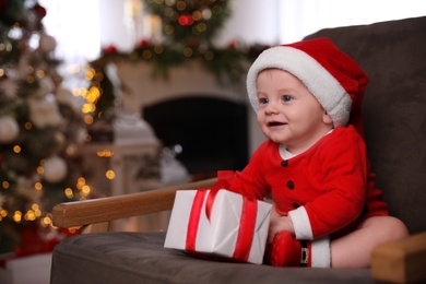 Cute little baby wearing Santa Claus suit with Christmas gift in armchair at home