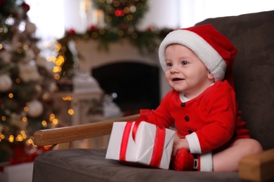 Photo of Cute little baby wearing Santa Claus suit with Christmas gift in armchair at home