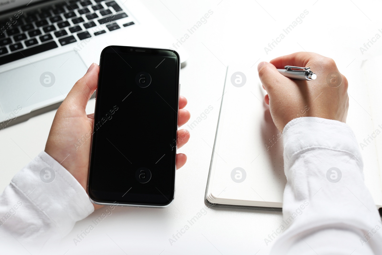 Photo of Woman with smartphone writing in notebook at white table, closeup