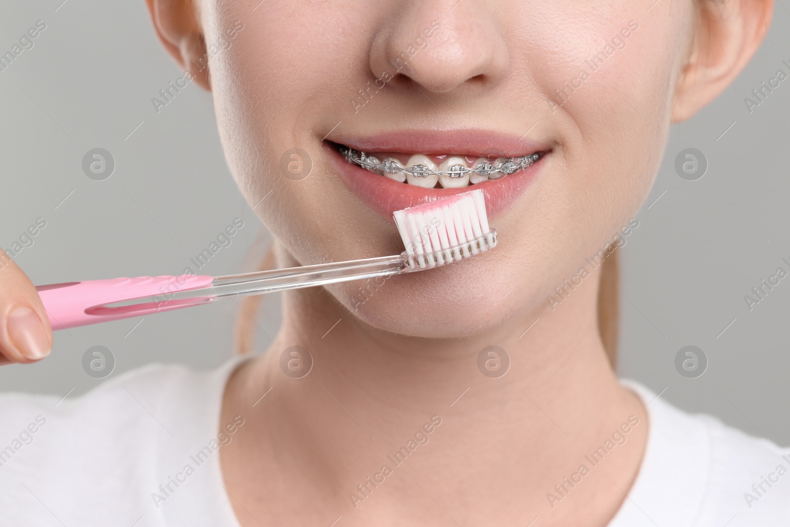 Photo of Smiling woman with dental braces cleaning teeth on grey background, closeup