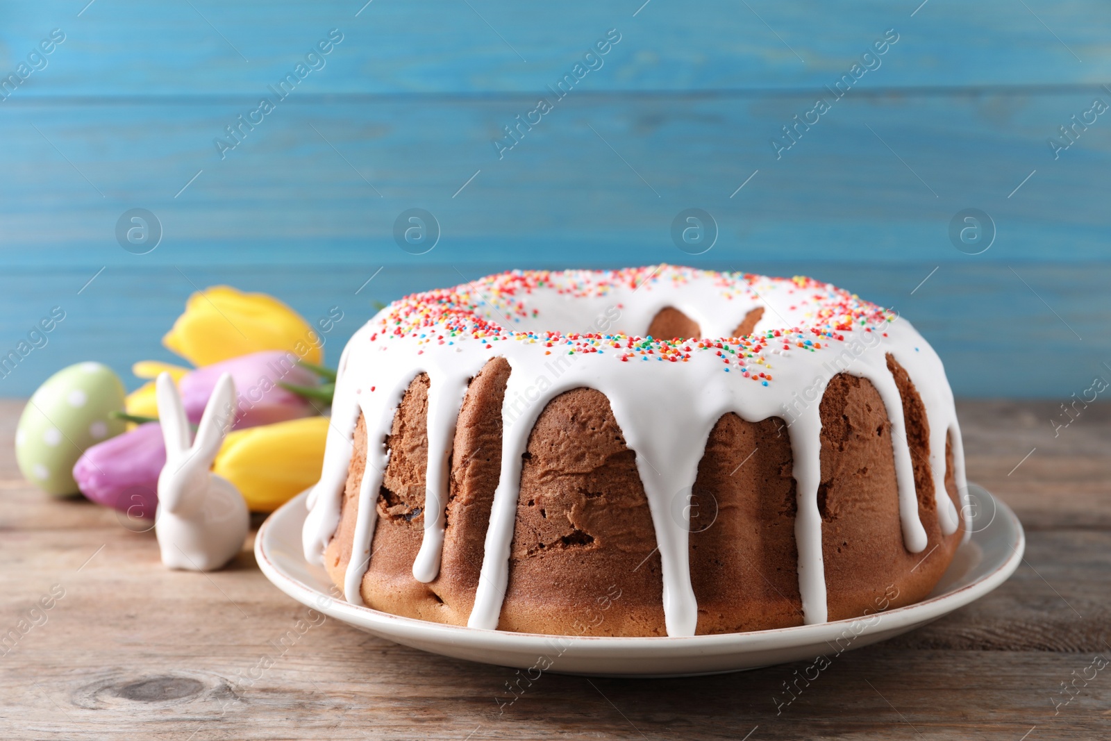 Photo of Glazed Easter cake with sprinkles, decorative bunny, painted egg and tulips on wooden table, closeup