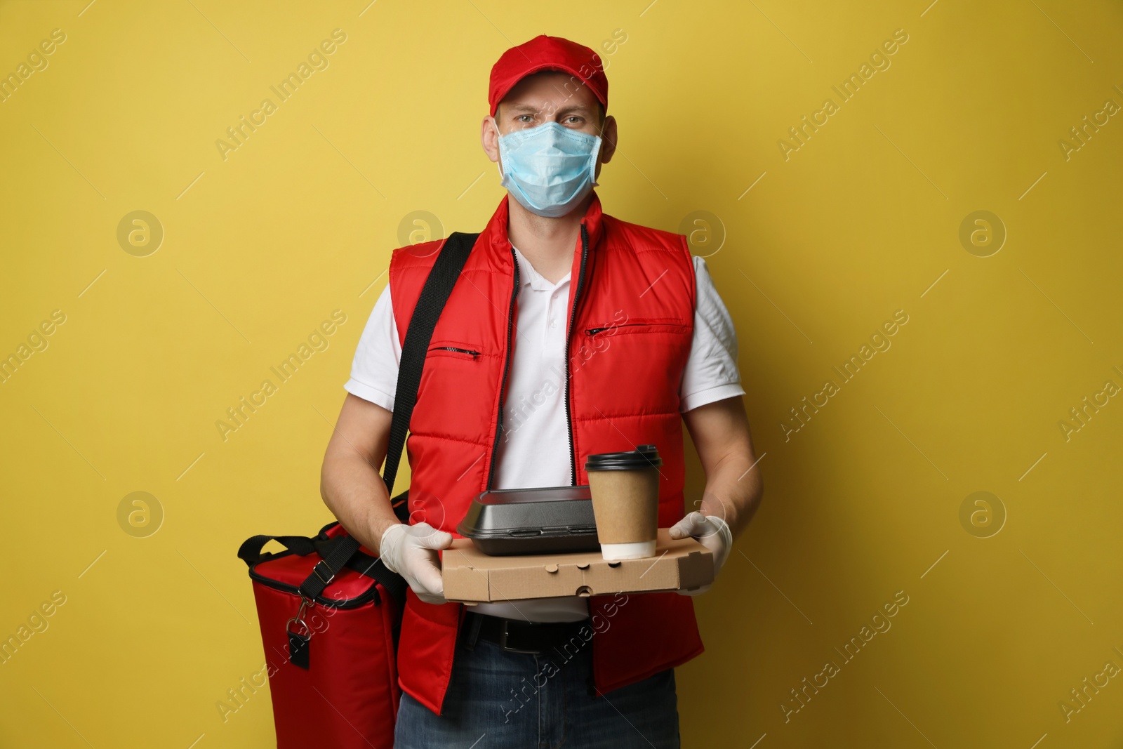 Photo of Courier in protective gloves and mask holding order on yellow background. Food delivery service during coronavirus quarantine