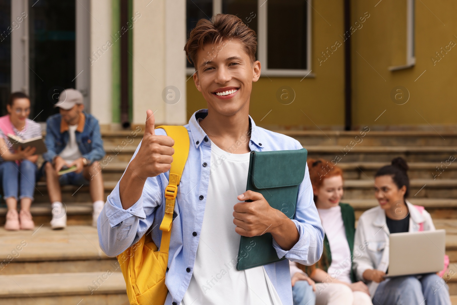 Photo of Students learning together on steps. Happy young man with tablet showing thumbs up outdoors, selective focus