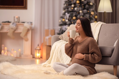 Happy pregnant woman with cup in room decorated for Christmas. Expecting baby