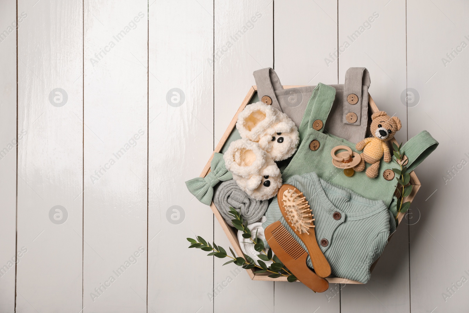 Photo of Children's clothes, shoes, toy bear and pacifier in crate on white wooden table, top view. Space for text