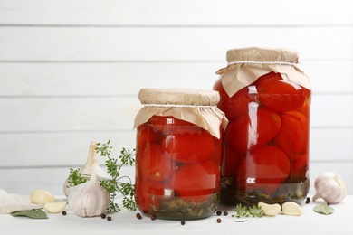 Glass jars of pickled tomatoes and ingredients on white wooden table