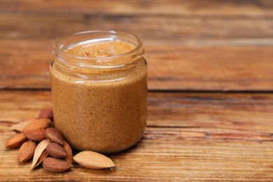 Tasty nut paste in jar and almonds on wooden table, closeup. Space for text