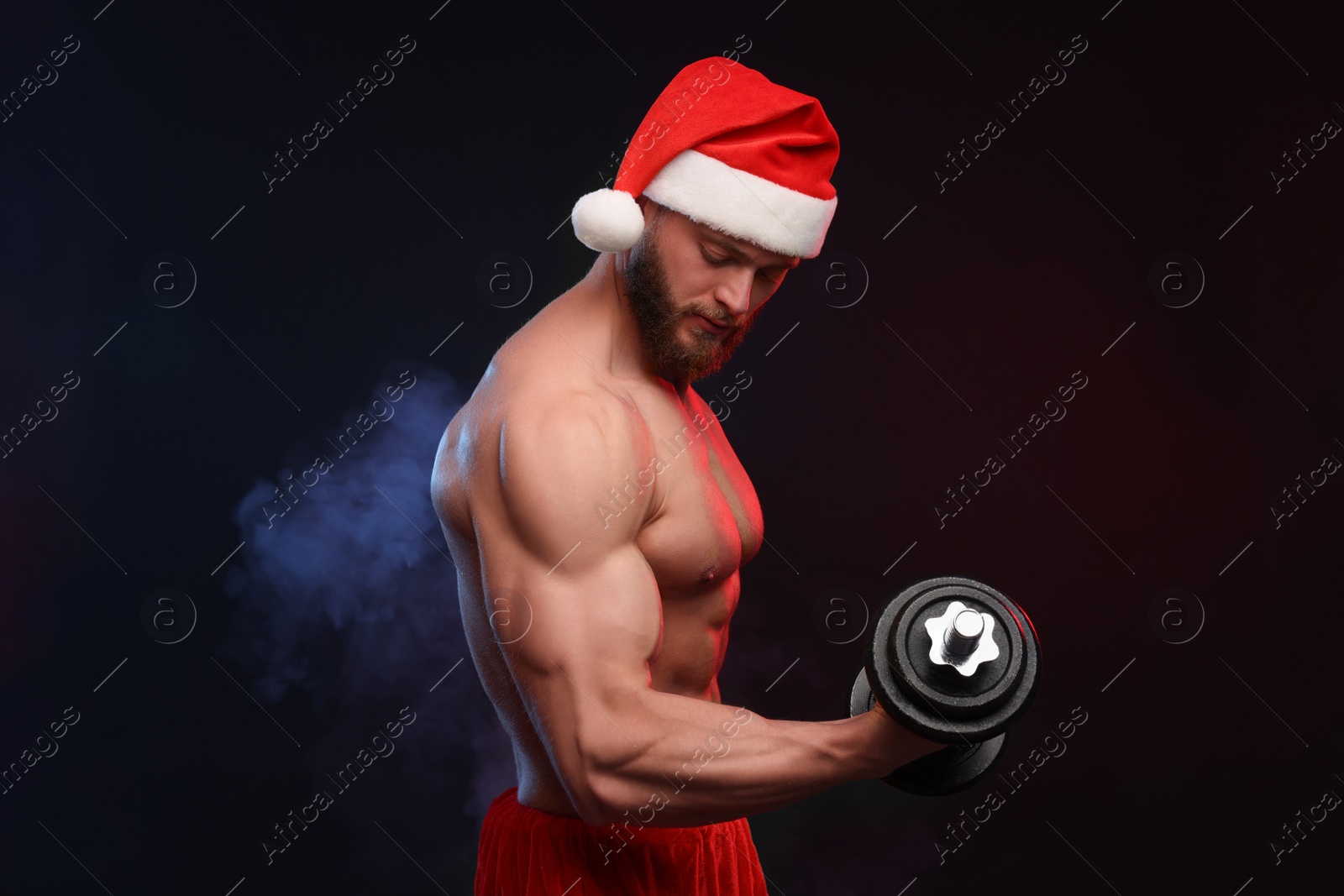 Photo of Muscular young man in Santa hat with dumbbell on black background