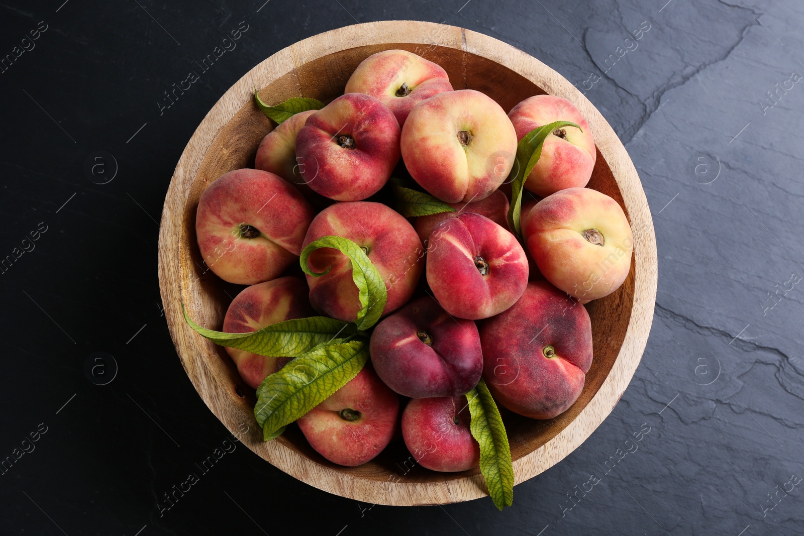 Photo of Fresh ripe donut peaches with leaves in bowl on dark table, top view