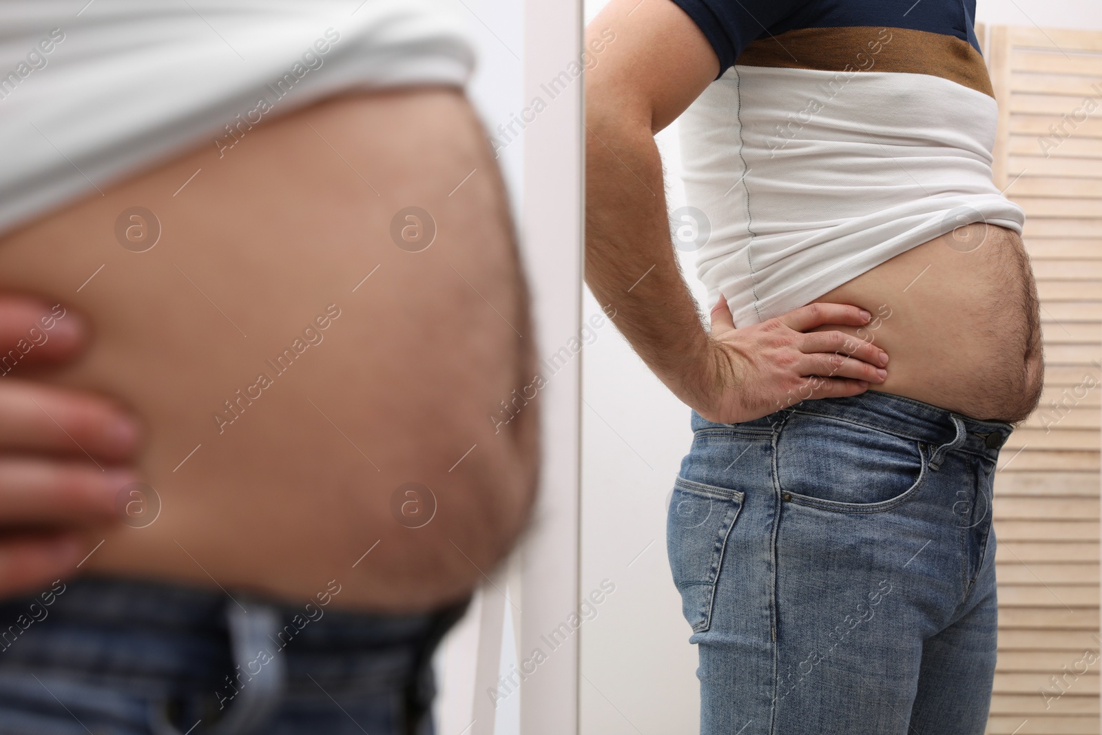 Photo of Man wearing tight t-shirt in front of mirror indoors, closeup. Overweight problem