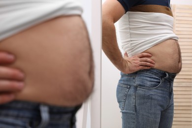 Photo of Man wearing tight t-shirt in front of mirror indoors, closeup. Overweight problem