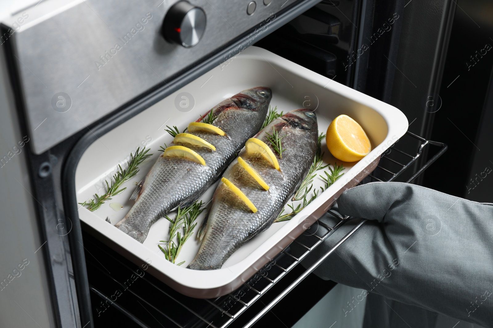 Photo of Woman putting baking tray with sea bass fish, lemon and rosemary into oven, closeup