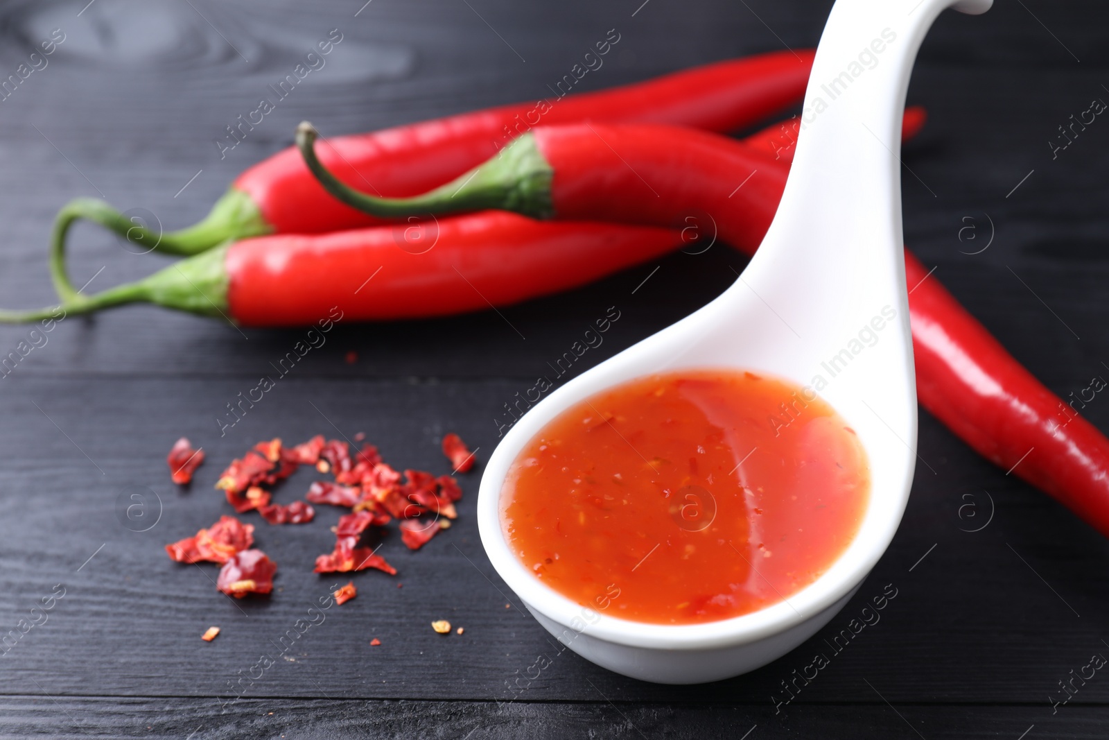 Photo of Spicy chili sauce in spoon and peppers on black wooden table, closeup