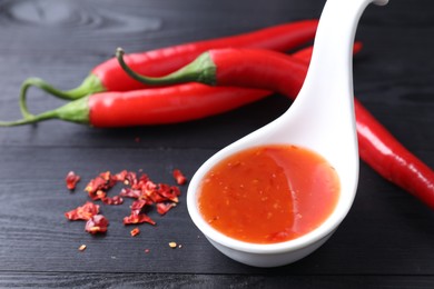 Spicy chili sauce in spoon and peppers on black wooden table, closeup