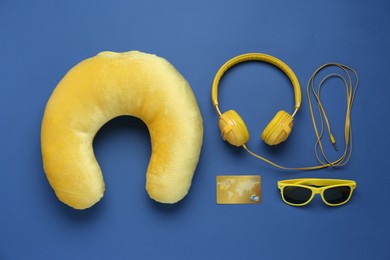 Photo of Yellow travel pillow, headphones, sunglasses and credit card on blue background, flat lay