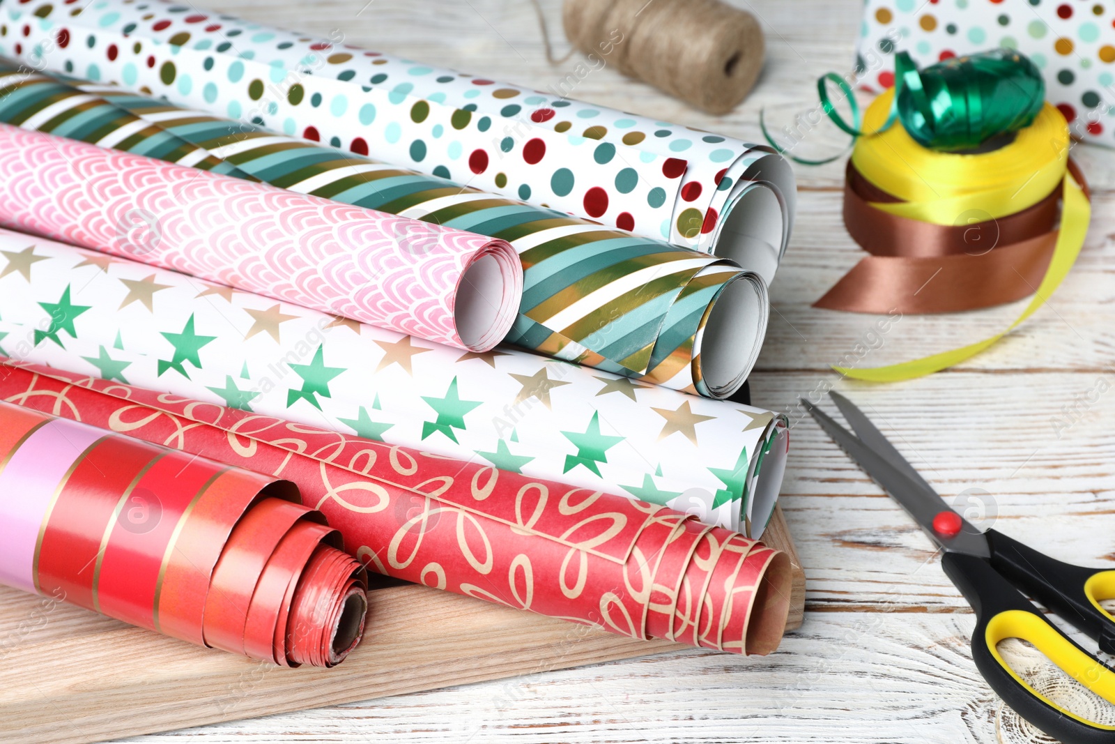 Photo of Different colorful wrapping paper rolls, scissors and ribbons on white wooden table