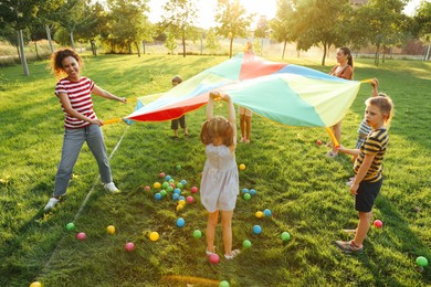 Photo of Group of children and teachers playing with rainbow playground parachute on green grass. Summer camp activity