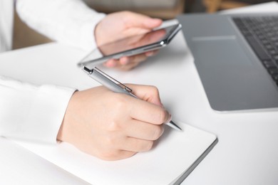 Photo of Woman with smartphone writing in notebook at white table in office, closeup