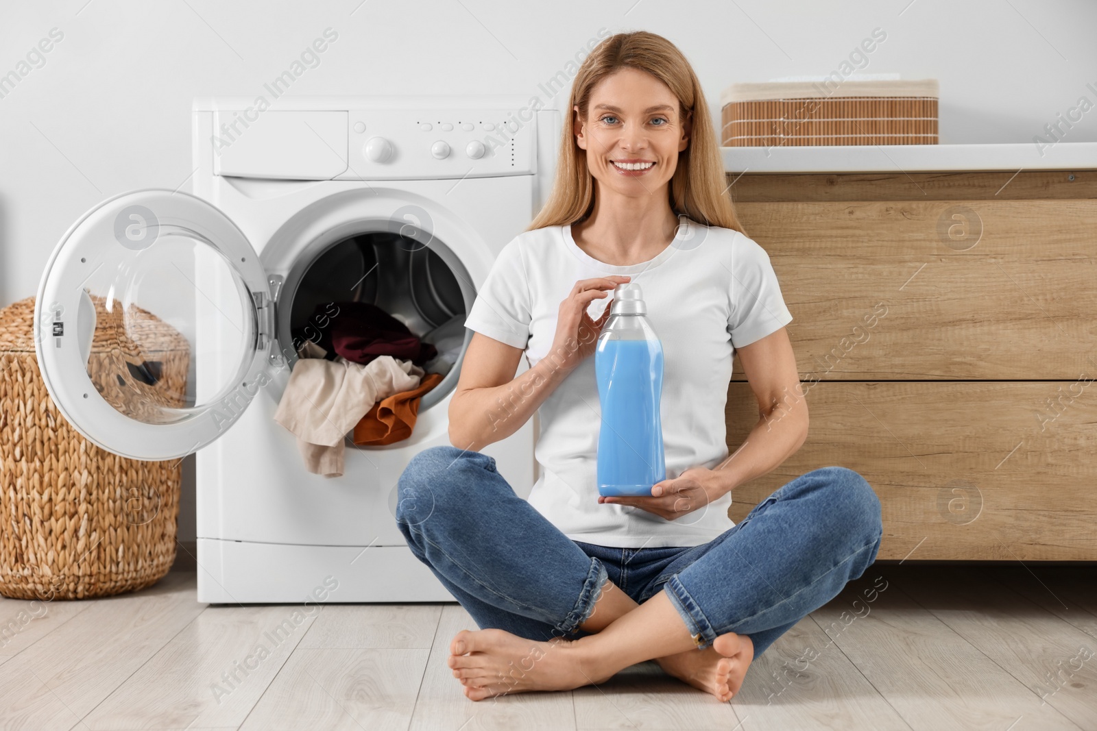 Photo of Woman sitting on floor near washing machine and holding fabric softener in bathroom