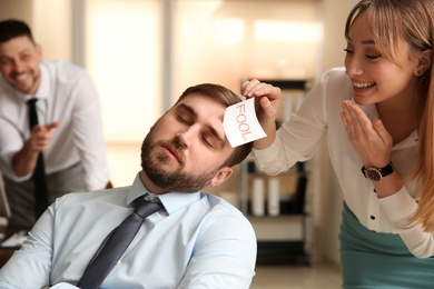 Photo of Young woman sticking note with word Fool to colleague's face in office. Funny joke