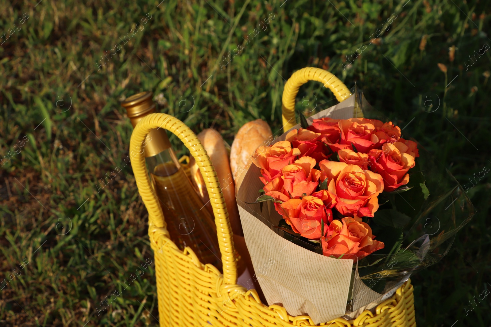 Photo of Yellow wicker bag with beautiful roses, bottle of wine and baguettes on green grass outdoors, closeup
