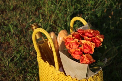 Yellow wicker bag with beautiful roses, bottle of wine and baguettes on green grass outdoors, closeup