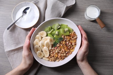 Woman holding bowl of tasty granola with banana and kiwi at grey wooden table, top view