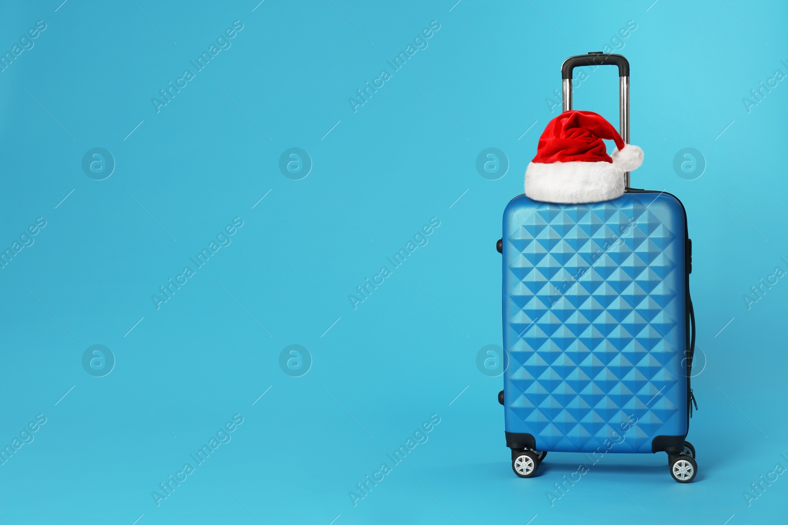 Image of Stylish suitcase with Santa Claus hat on blue background, space for text. Christmas vacation 