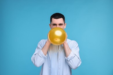 Photo of Young man inflating golden balloon on light blue background