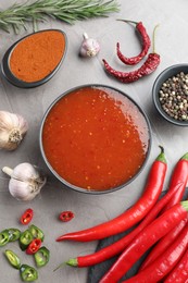 Photo of Spicy chili sauce in bowl and ingredients on grey table, flat lay