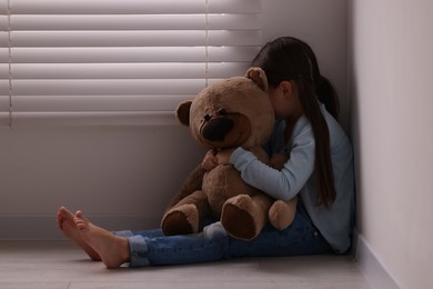 Child abuse. Upset little girl with teddy bear sitting on floor near light wall indoors