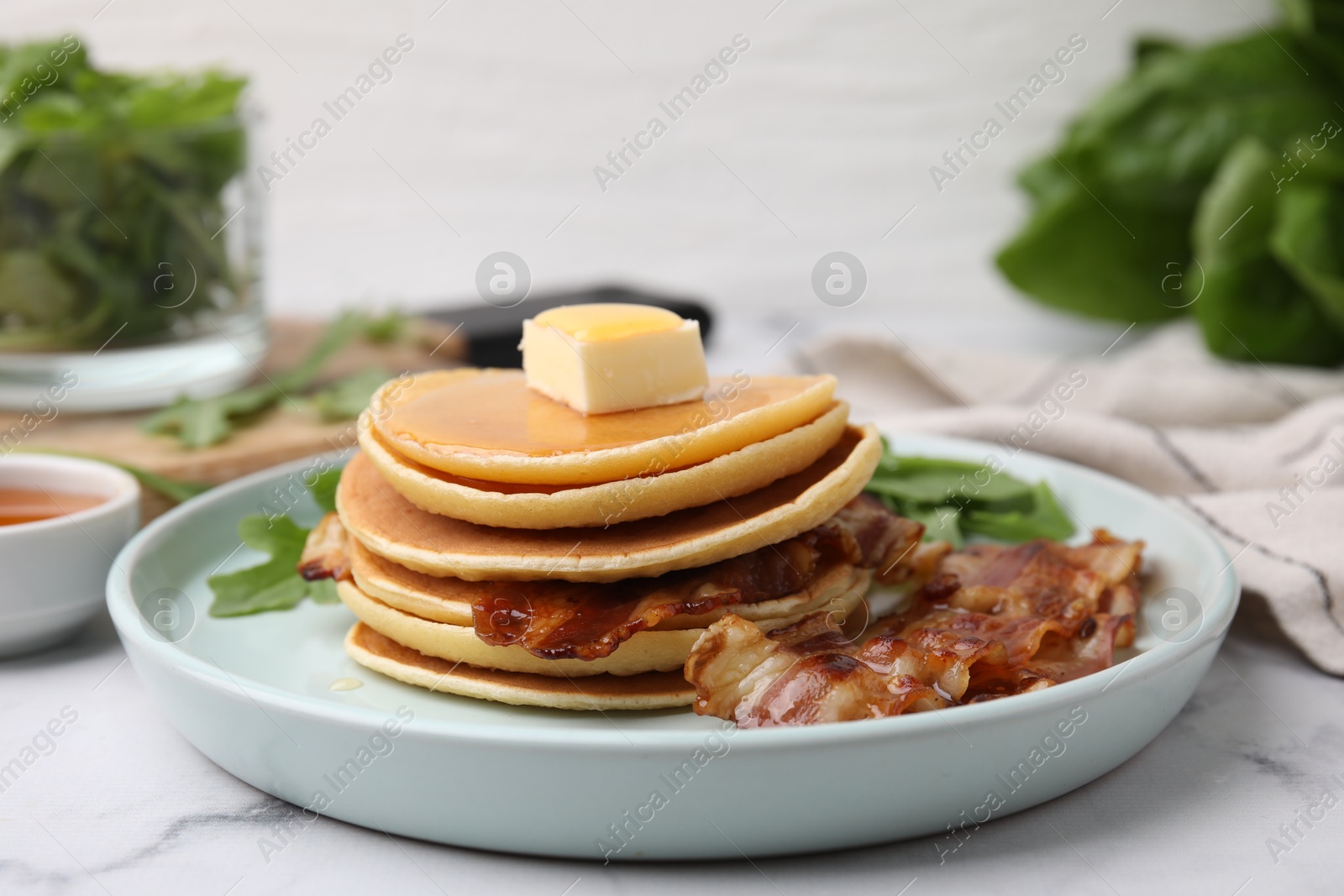 Photo of Tasty pancakes with butter, fried bacon and fresh arugula on white marble table