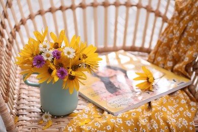 Photo of Beautiful bright flowers in light blue cup near magazine and fabric on rattan armchair. Space for text