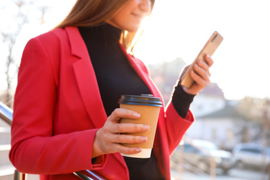 Photo of Businesswoman with cup of coffee and smartphone on city street in morning, closeup