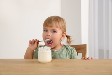 Cute little child eating tasty yogurt with spoon at wooden table indoors