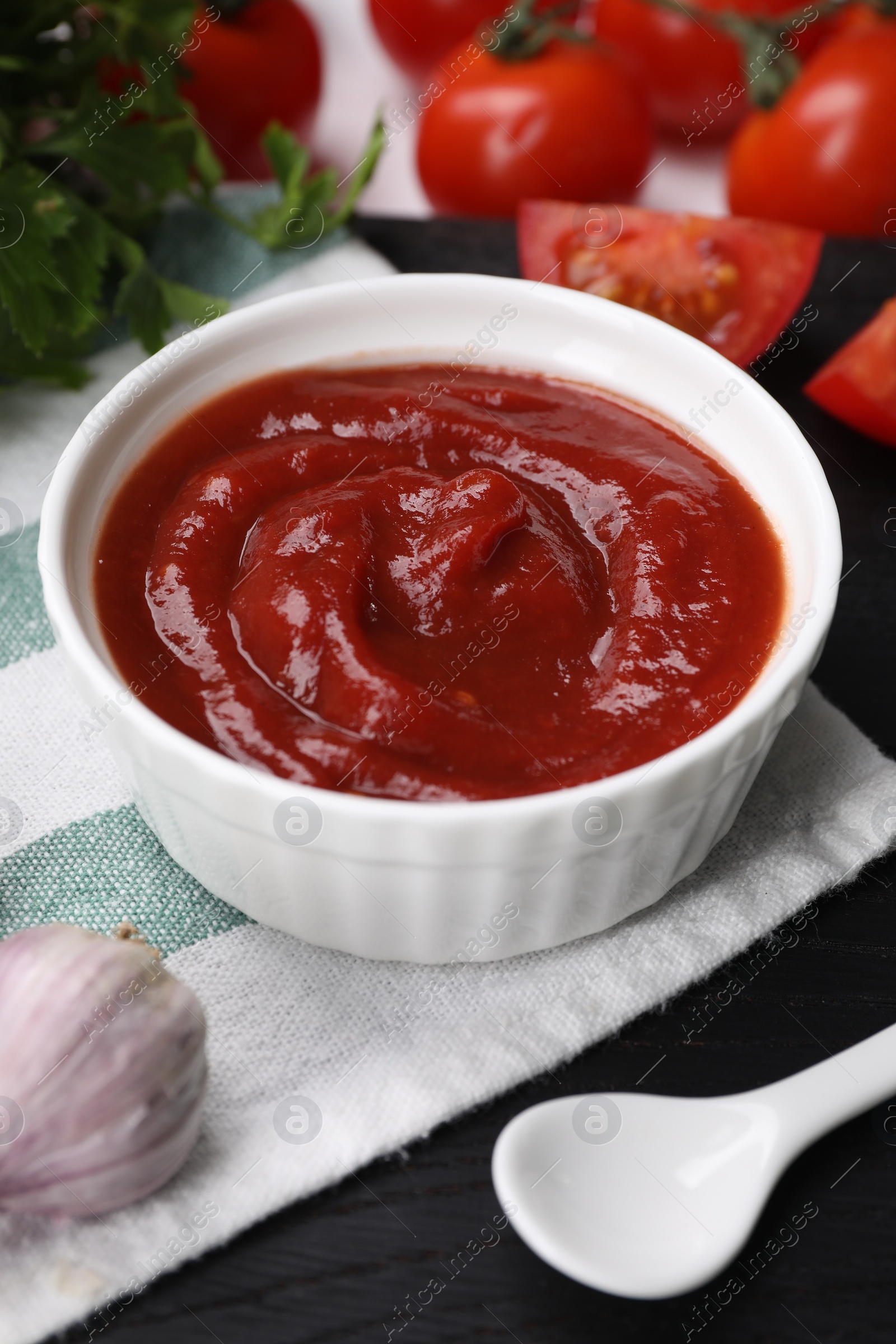 Photo of Organic ketchup in bowl, fresh tomatoes and garlic on black table, closeup. Tomato sauce