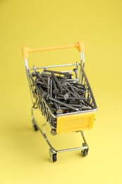Photo of Metal nails in shopping cart on yellow background, closeup