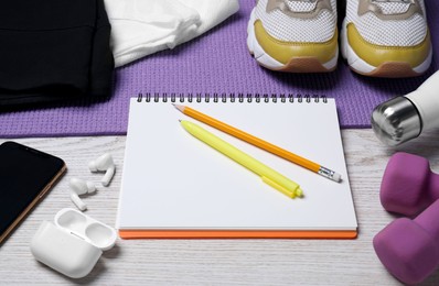 Photo of Notebook, wireless earphones, clothes and sports equipment on white wooden table. Personal training