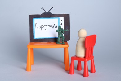 Photo of Hybrid warfare concept. Human figure watching TV propaganda at gunpoint by toy soldier on light grey background