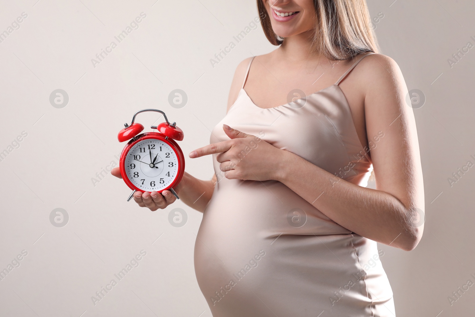 Photo of Young pregnant woman pointing at alarm clock near her belly on beige background, closeup. Time to give birth