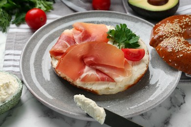 Photo of Delicious bagel with cream cheese, jamon, tomato and parsley on white marble table, closeup