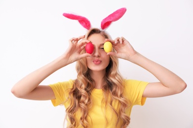 Photo of Beautiful young woman in bunny ears headband holding Easter eggs near eyes on light background