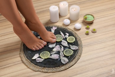 Photo of Woman soaking her feet in plate with water, flower petals and lime slices on wooden floor, closeup. Pedicure procedure