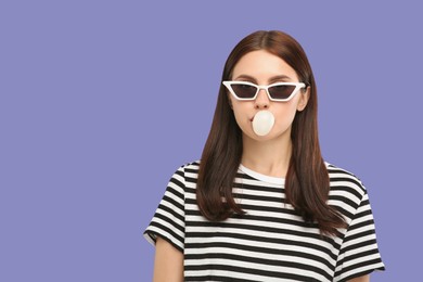 Beautiful woman in sunglasses blowing bubble gum on light purple background, space for text