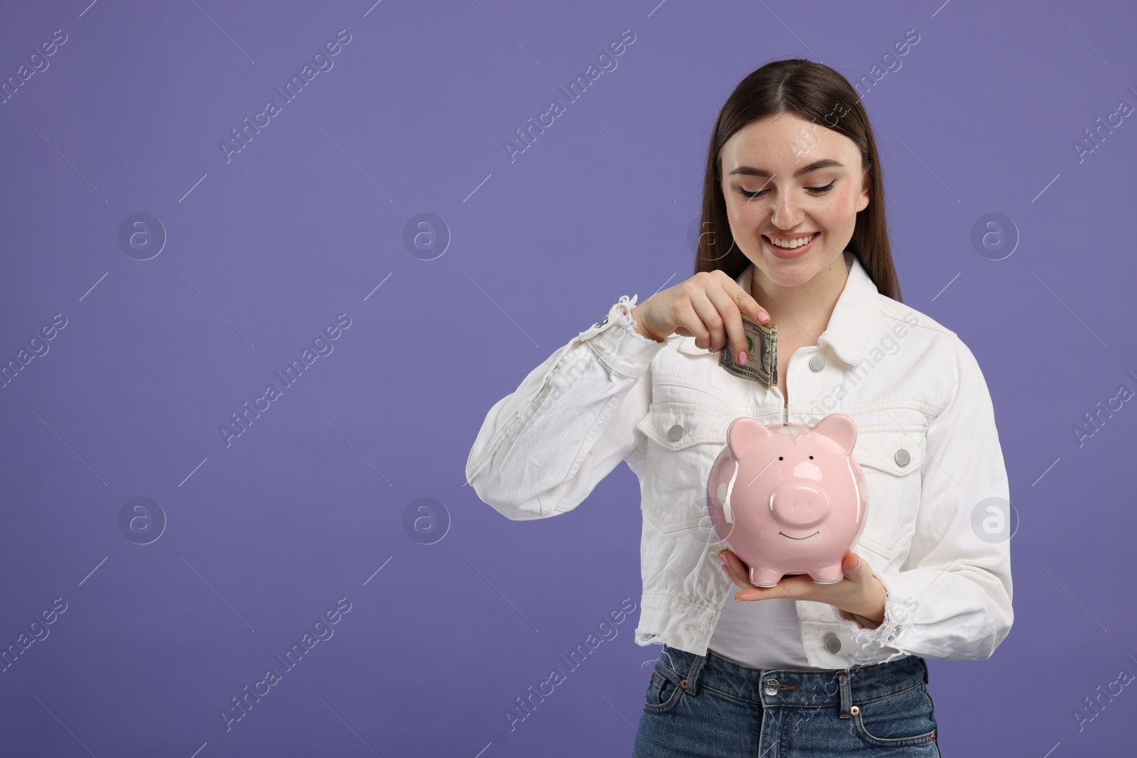 Photo of Happy woman putting dollar banknote into piggy bank on purple background, space for text