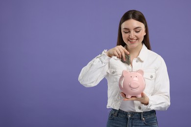 Happy woman putting dollar banknote into piggy bank on purple background, space for text