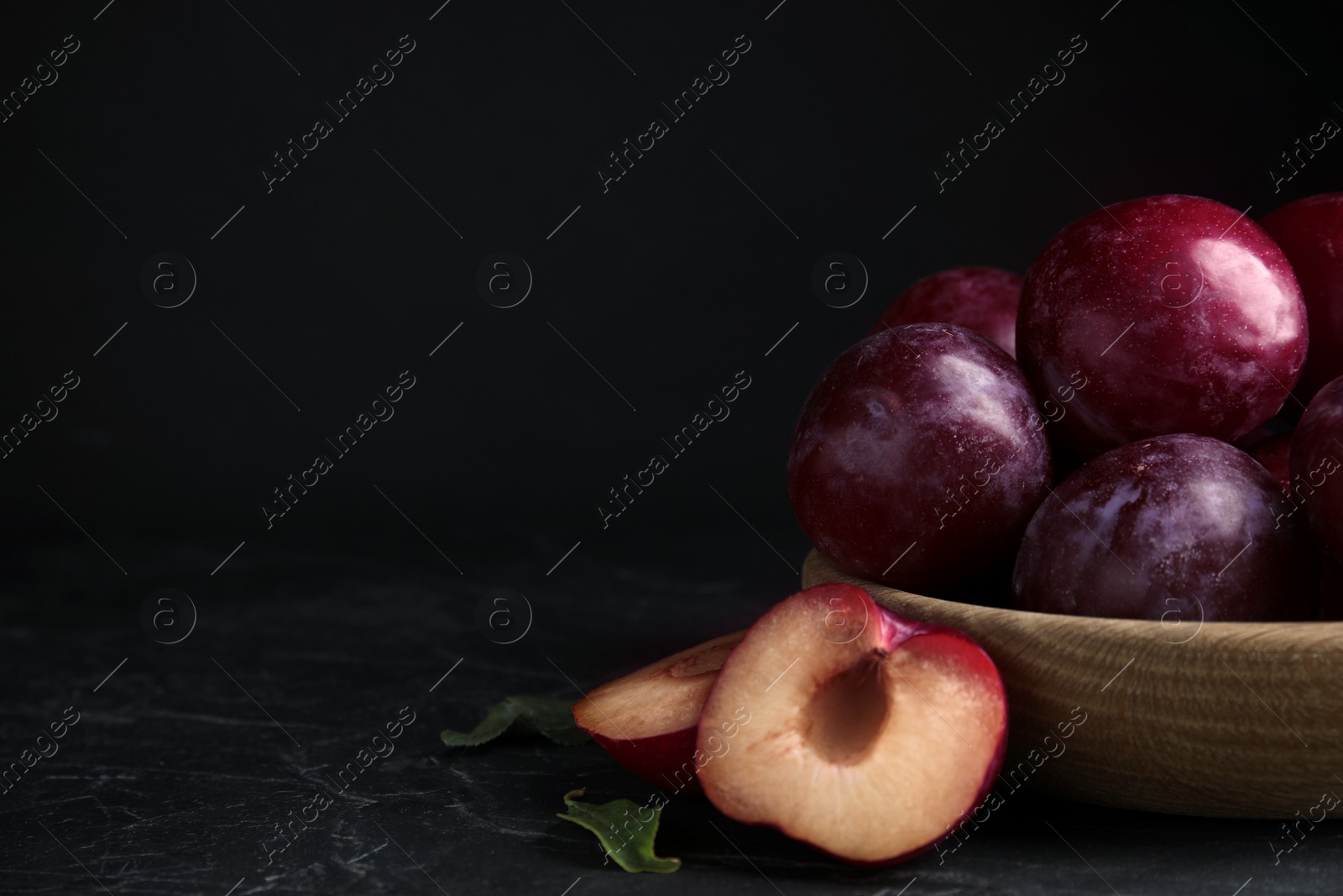 Photo of Delicious ripe plums in wooden bowl on black table. Space for text
